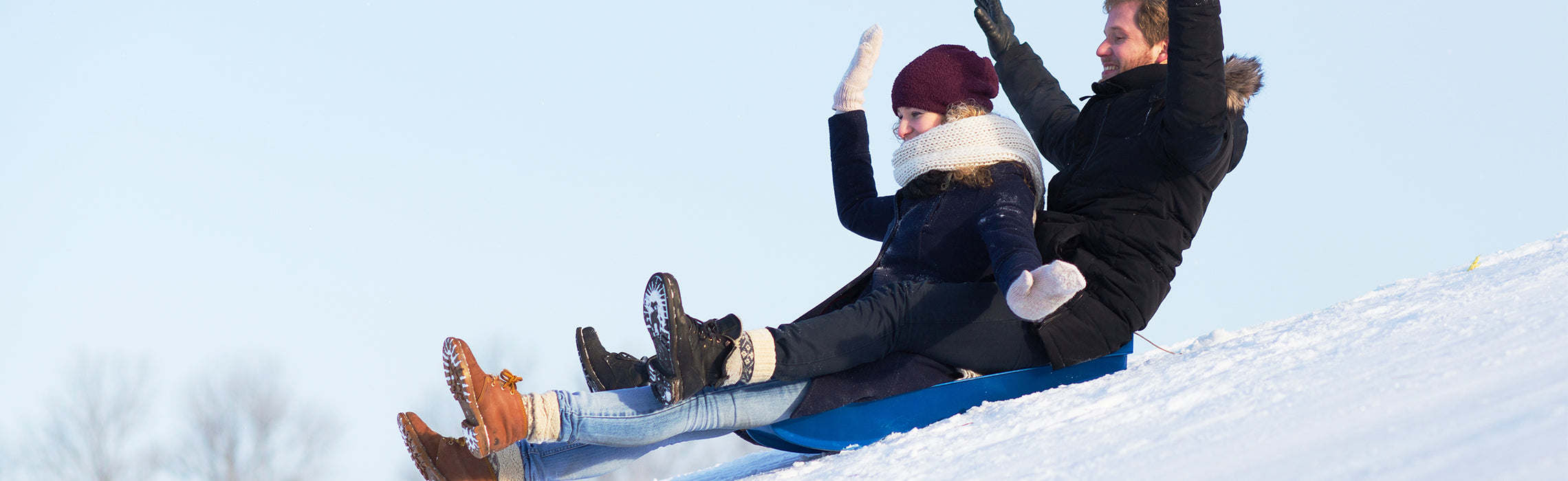 Two people bundled in winter clothing sled down a snowy hill on a blue sled, lifting their arms as they glide across a bright, snowy landscape. No visible text or branding.
