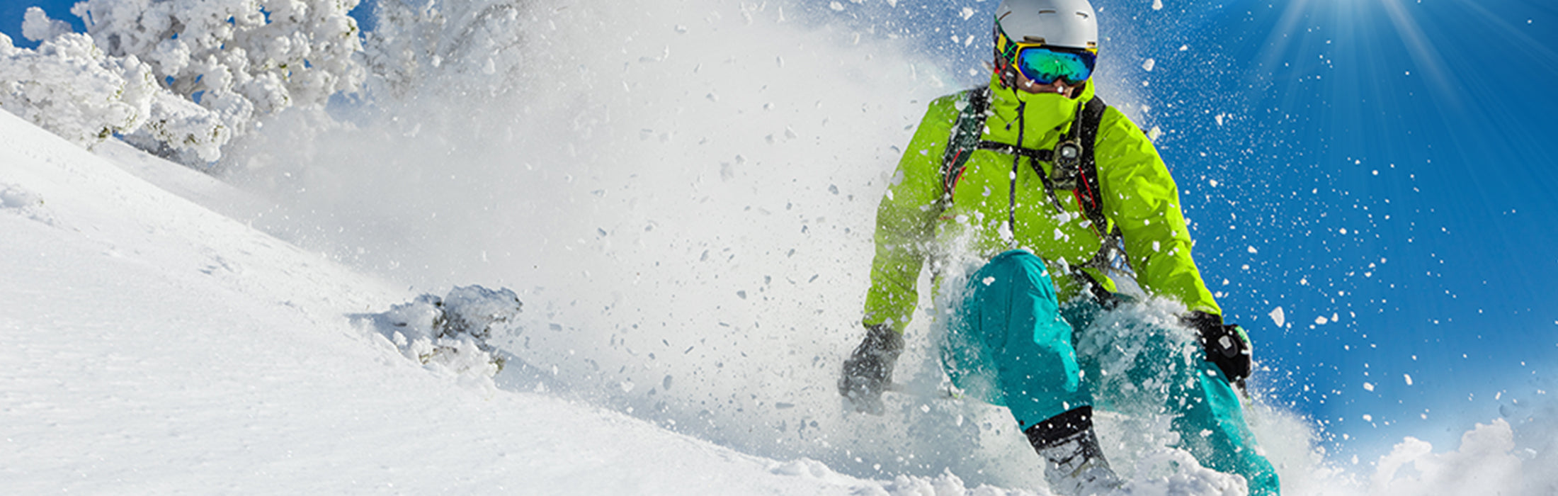 Person wearing a helmet, goggles, and bright outdoor gear skiing down a snowy mountain, kicking up powder against a clear blue sky, capturing winter sports action. No visible text or branding.