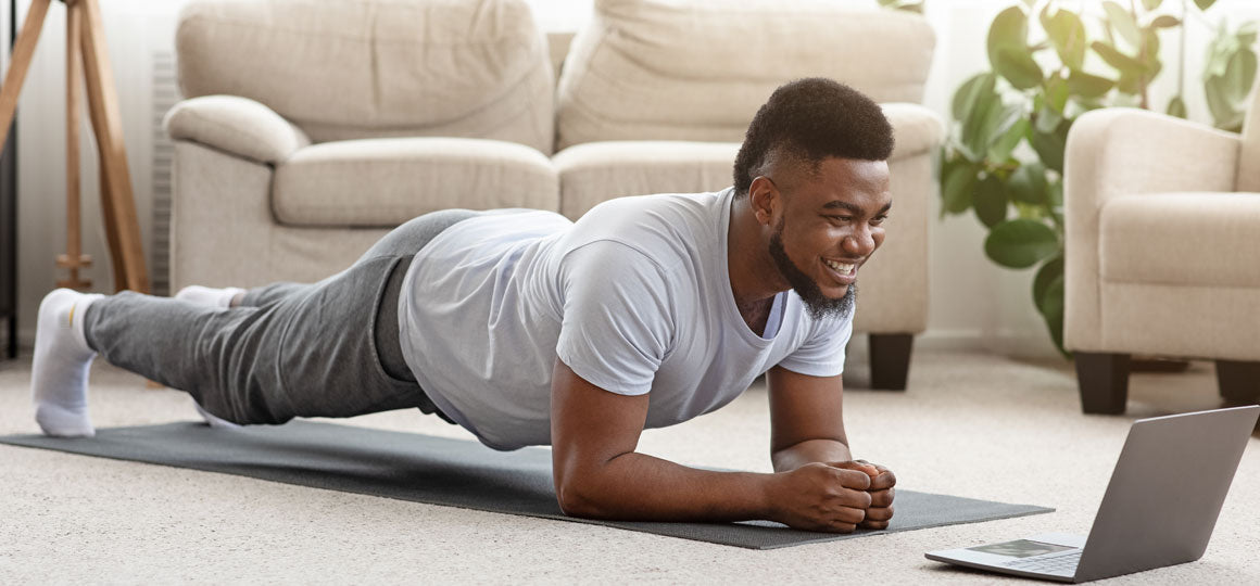 Person holding a forearm plank position on an exercise mat while watching a laptop in a bright living room with a sofa and houseplant in the background. No visible text or branding.