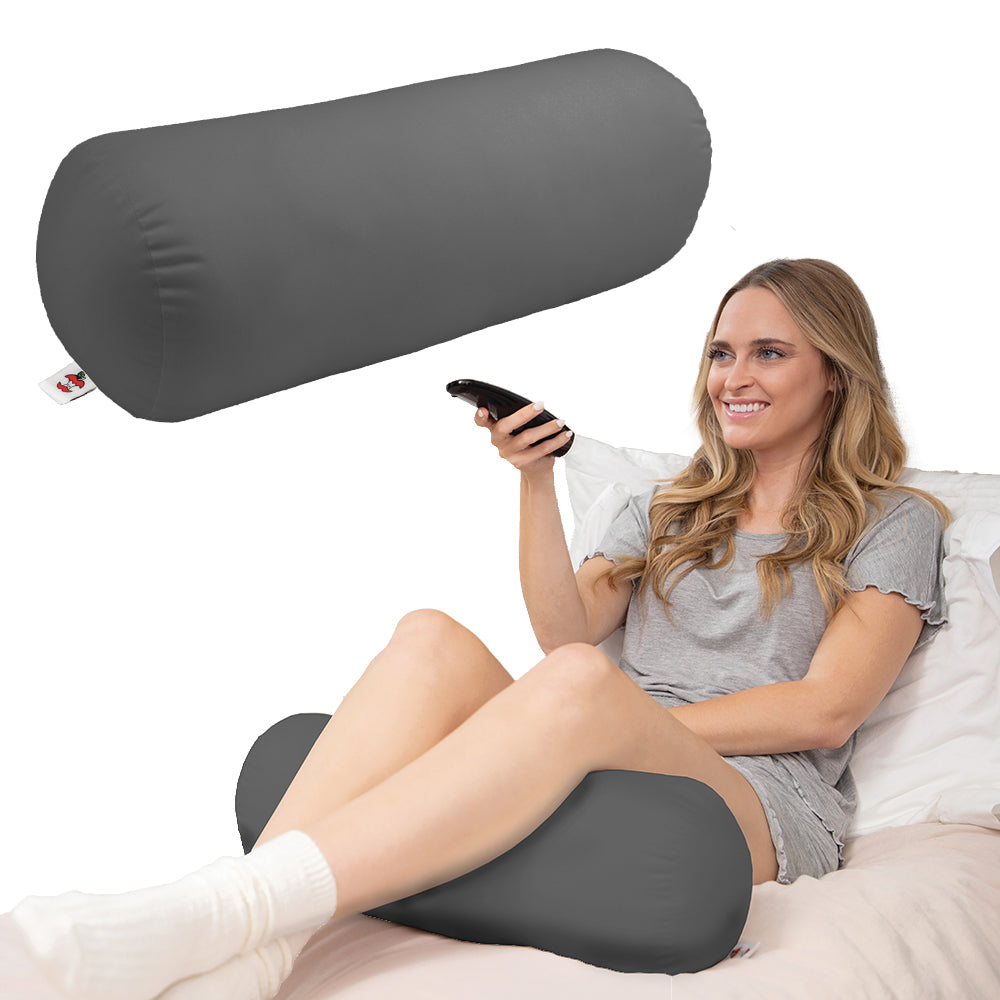 Core Products Jackson Roll Positioning Support Pillow in gray shown supporting a woman sitting comfortably in bed. Cylindrical fiber-filled pillow provides versatile support for legs, knees, and back, promoting proper alignment, relaxation, and everyday comfort.