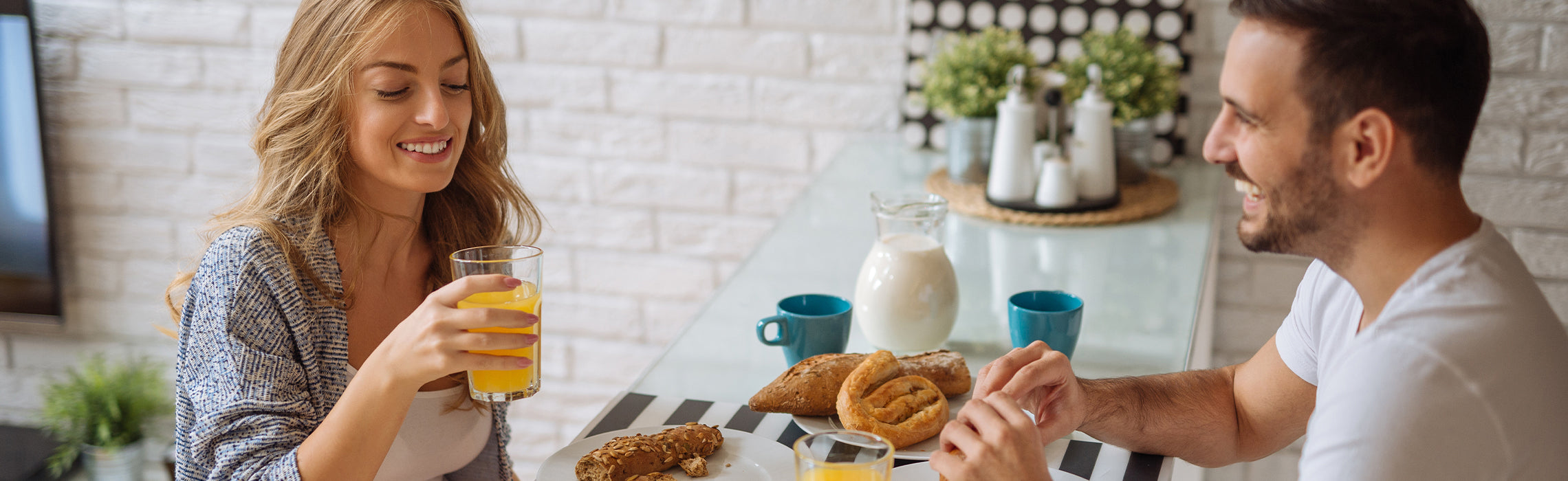 Two people sitting at a table enjoying breakfast with bread, pastries, and glasses of orange juice, with a pitcher of milk and cups nearby, representing healthy eating, morning routine, and home lifestyle.