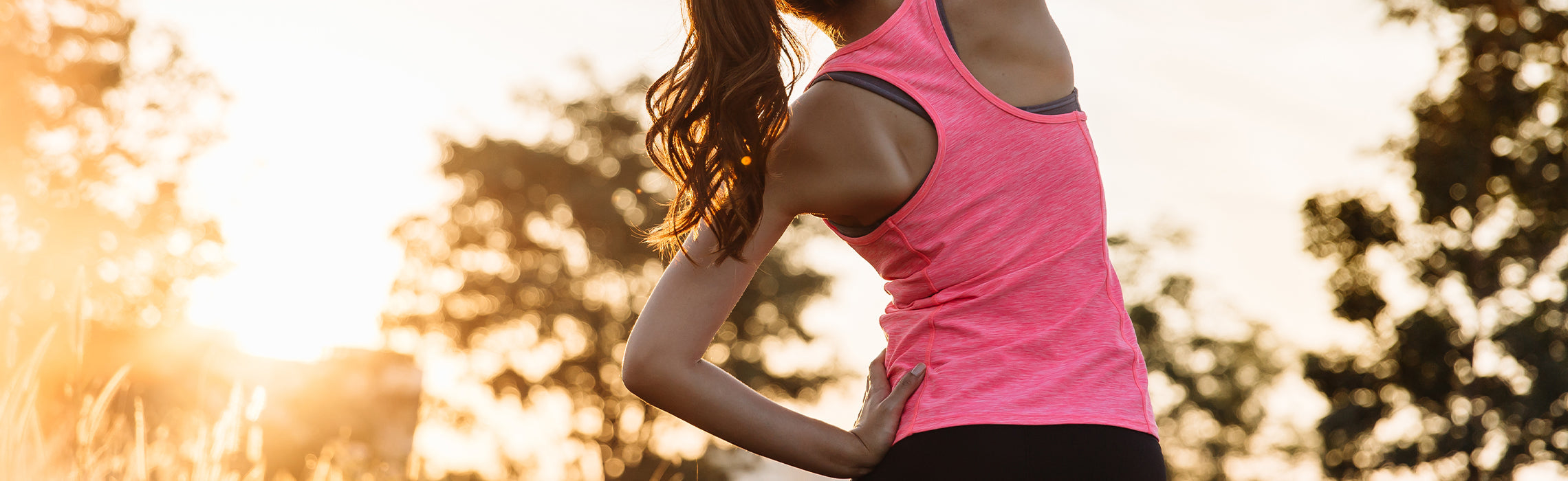 Person in a pink athletic top stretching the side of the torso outdoors at sunset with a hand on the hip, representing warm‑up exercise, flexibility, and outdoor fitness.