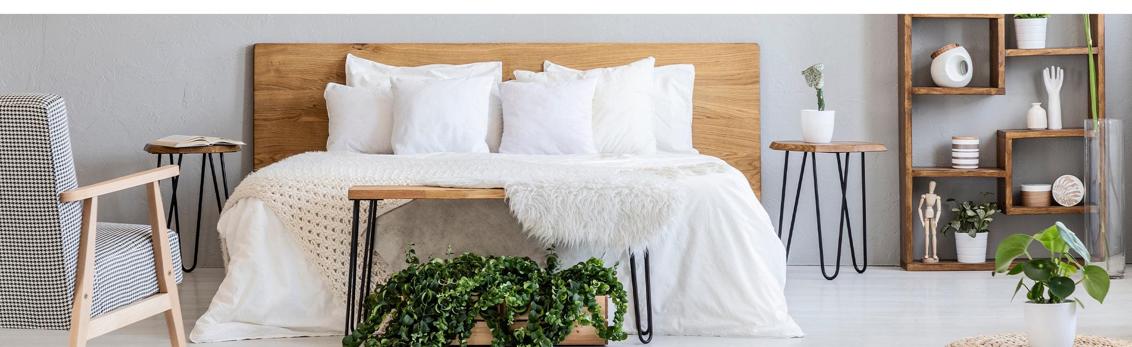 Bright, minimalist bedroom with a wooden bed frame, white bedding and pillows, a knit throw, and green plants, alongside modern side tables, a chair, and decorative shelving, representing cozy home décor and comfortable sleep space.