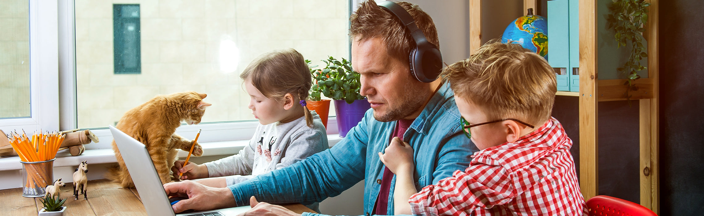 Person using a laptop at a desk while wearing headphones, with two children nearby and a cat sitting beside the keyboard, representing working from home, multitasking, and family life.