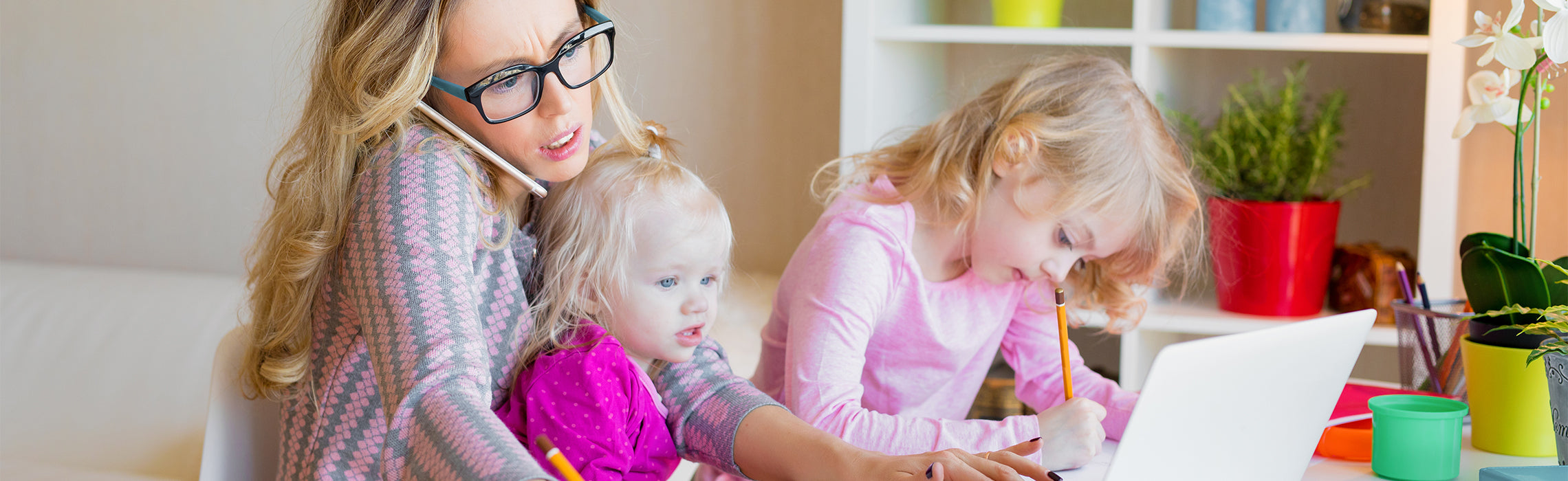 Person holding a phone while working at a desk with a laptop, supporting a young child on their lap as another child writes with a pencil nearby, illustrating multitasking at home. No visible text or branding.