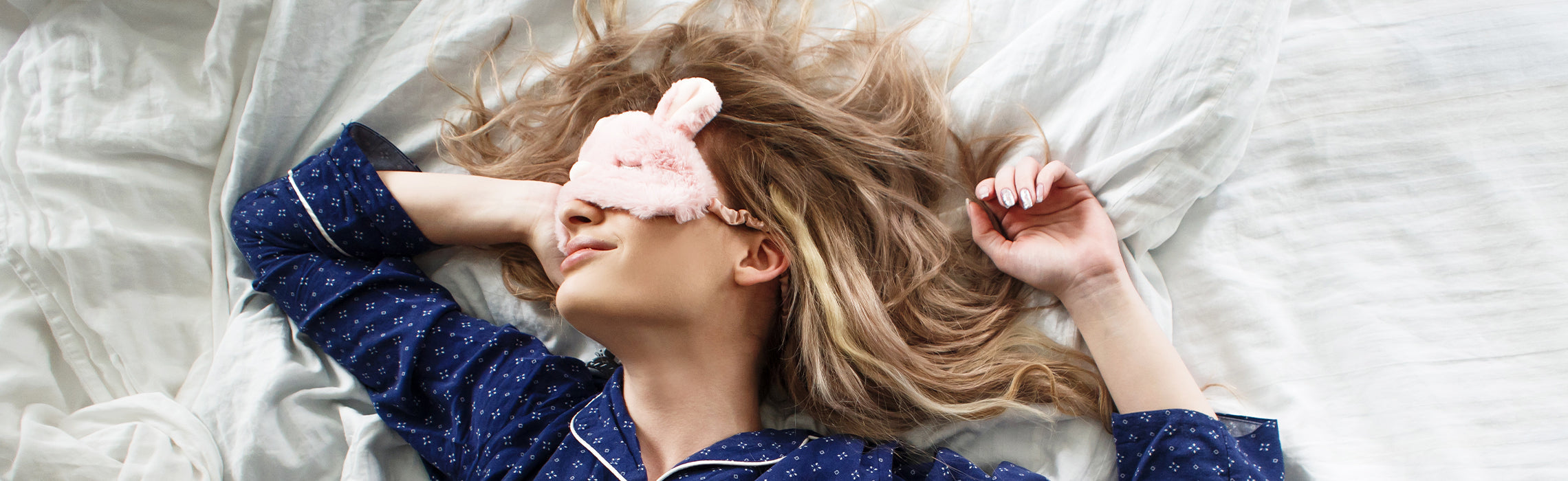 Woman sleeping comfortably in bed wearing a pink sleep mask and blue pajamas, resting on white bedding.