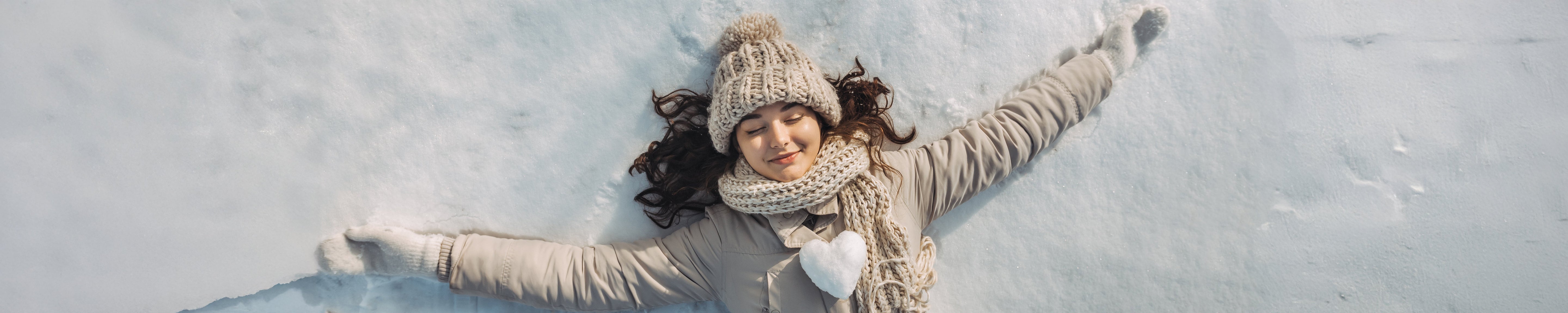 Person lying on snow with arms outstretched, wearing a knit hat, scarf, gloves, and winter coat, creating a snow angel on a bright winter day. No visible text or branding.