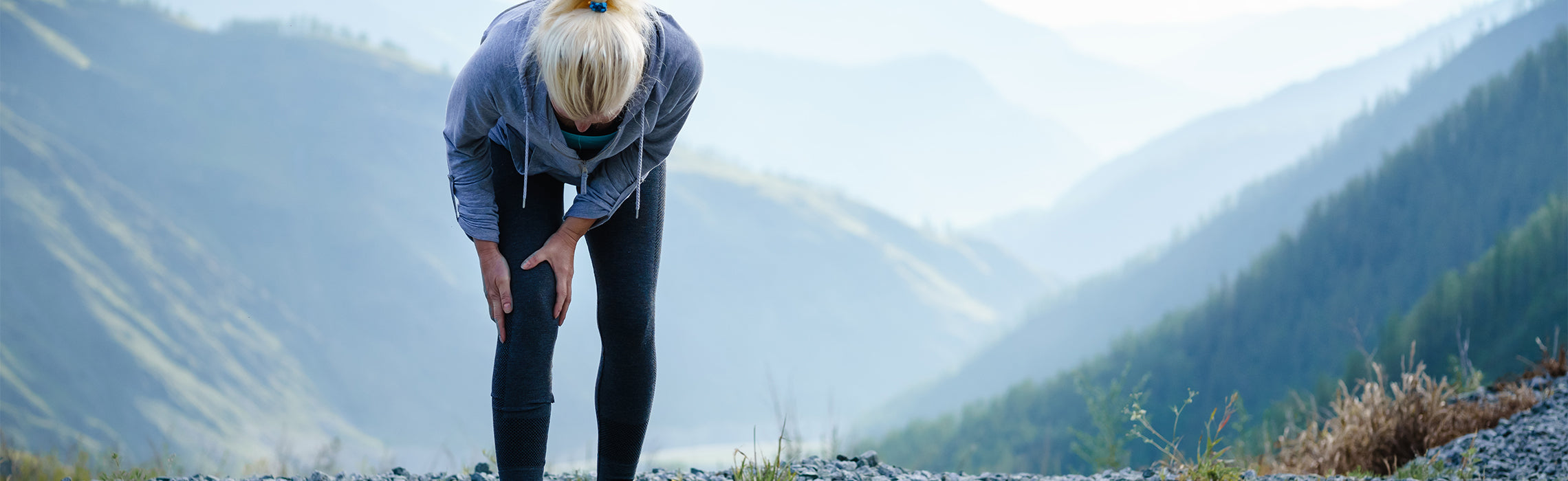 Person standing outdoors on a rocky path holding one knee with both hands, with mountains in the background, representing knee pain, hiking injury, and joint strain during exercise.