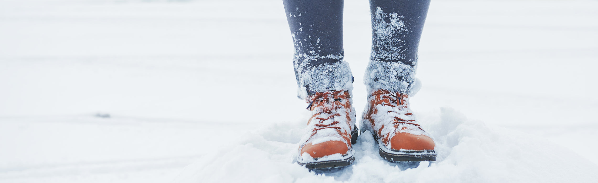 Close-up of orange winter boots standing in deep snow with pant legs dusted in snow, representing cold weather conditions, winter footwear, and outdoor activity in snowy terrain.