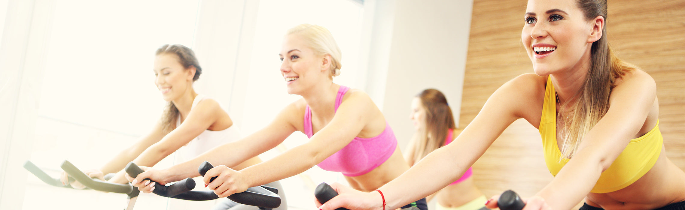 Three people using stationary bikes in a bright fitness studio, wearing athletic clothing, representing indoor cycling, group exercise, and cardio workout training.