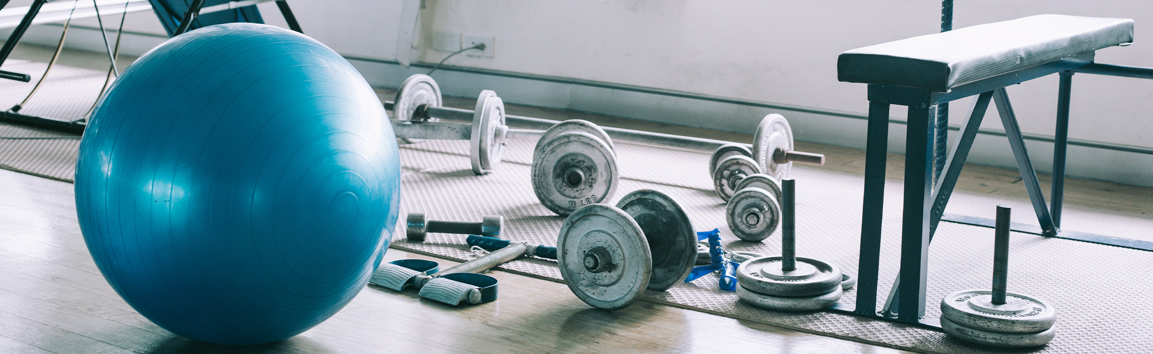 Blue exercise ball, dumbbells, weight plates, and resistance bands scattered on a gym floor next to a workout bench, representing strength training equipment, home gym setup, and fitness routines.