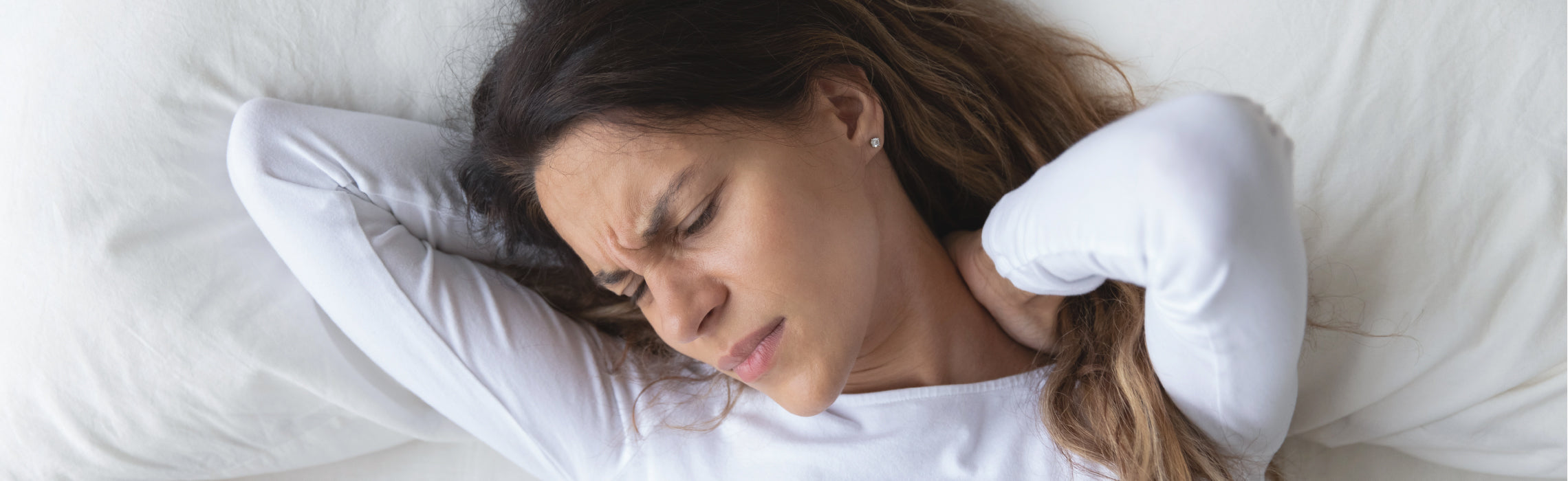 Person lying on a bed holding the neck with both hands, indicating neck pain or stiffness, on white bedding in a bright bedroom setting.