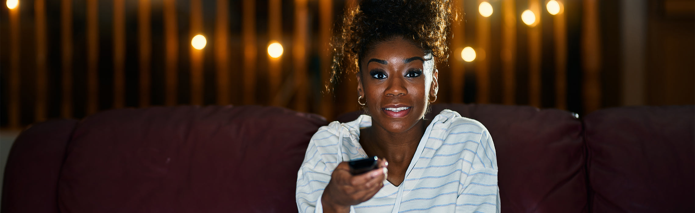 Person sitting on a couch holding a TV remote, facing forward in a softly lit living room with warm lights in the background. No visible text or branding.