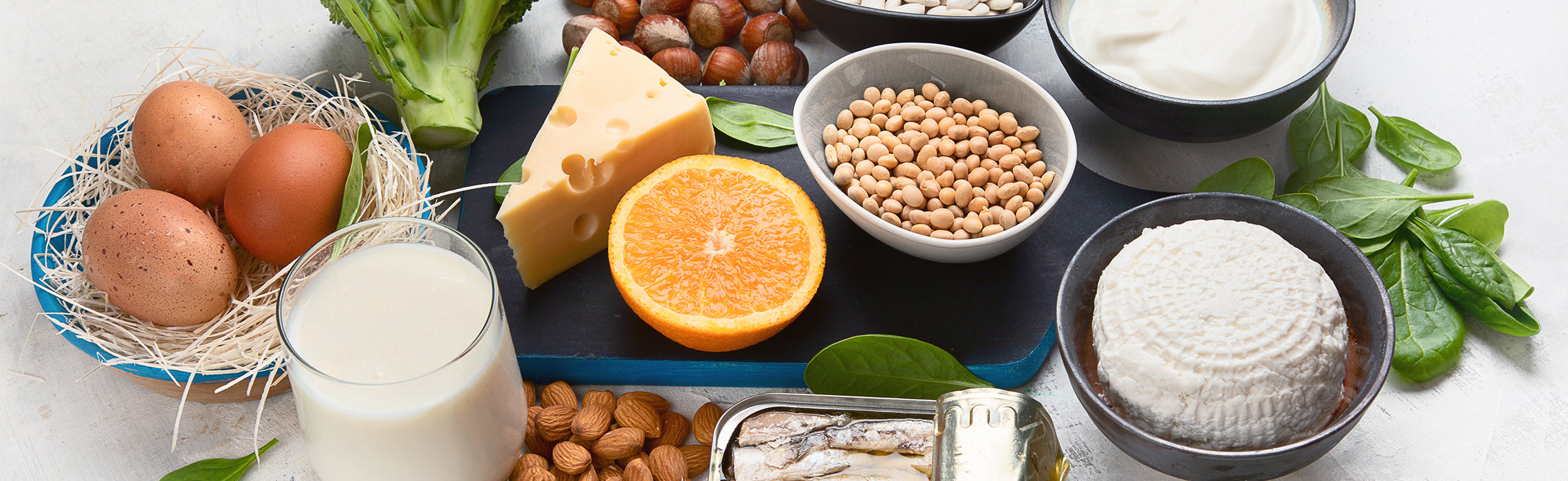 Assortment of nutrient-rich foods including eggs, milk, cheese, yogurt, nuts, beans, fish, broccoli, spinach, and orange on a tabletop, representing a balanced, high-protein and calcium-rich healthy diet.