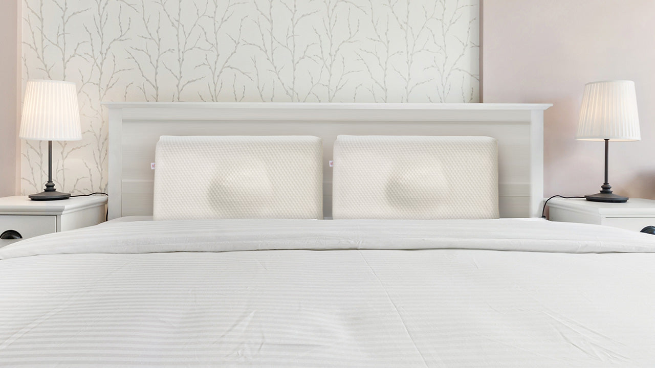 Neatly made bed with white bedding and two memory foam pillows against a white headboard, flanked by matching lamps in a minimalist bedroom. No visible text or branding.