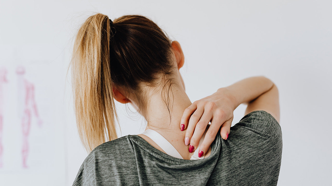 Close-up of a person touching the back of the neck with one hand, hair in a ponytail, suggesting neck tension or discomfort against a light indoor background. No visible text or branding.