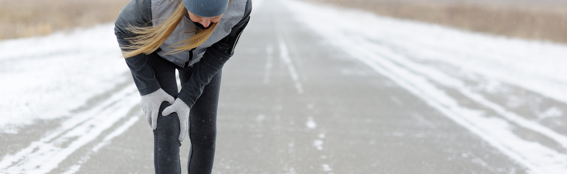Person outdoors on a snowy road bending forward while holding one knee with both gloved hands, wearing winter running gear, representing knee pain, injury, and cold‑weather exercise strain.