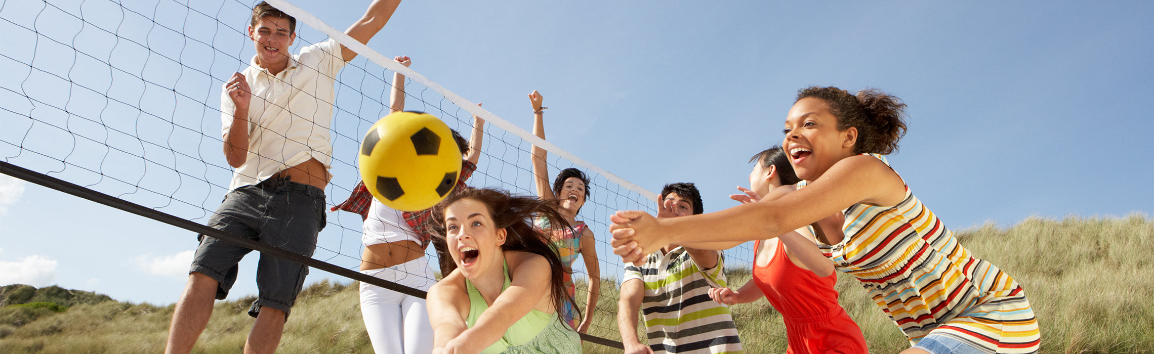 Group of people playing beach volleyball outdoors, jumping and reaching for a yellow ball over a net on a sunny day, representing team sports, active lifestyle, and outdoor recreation.
