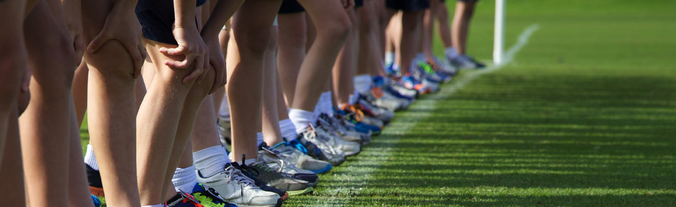 Close-up of athletes lined up on a grass field wearing running shoes, with one person holding a knee, representing knee pain, sports injury, and team fitness training.