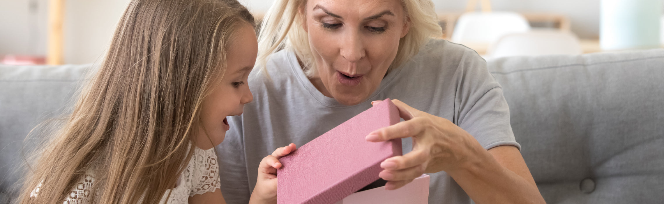 Close-up of an older adult and a child sitting on a couch opening a small pink gift box together, revealing a surprise, representing family bonding, gifting, and special moments.