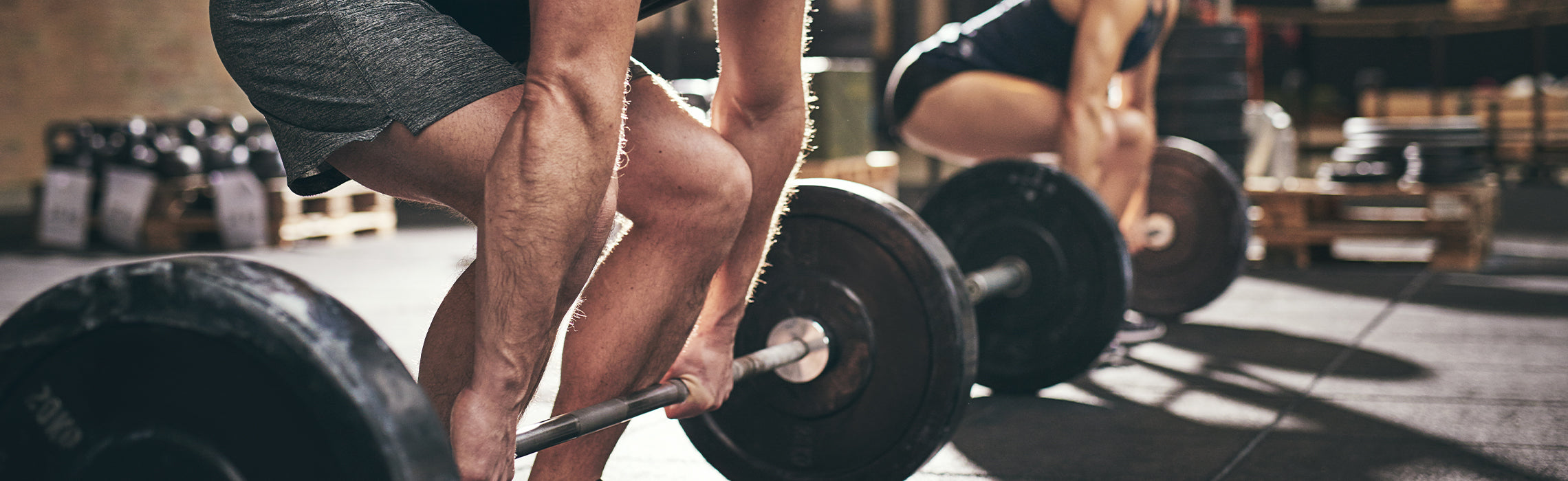 Close-up of a person gripping a barbell while lifting weights in a gym, with another person in the background also exercising, representing strength training, fitness workouts, and weightlifting.