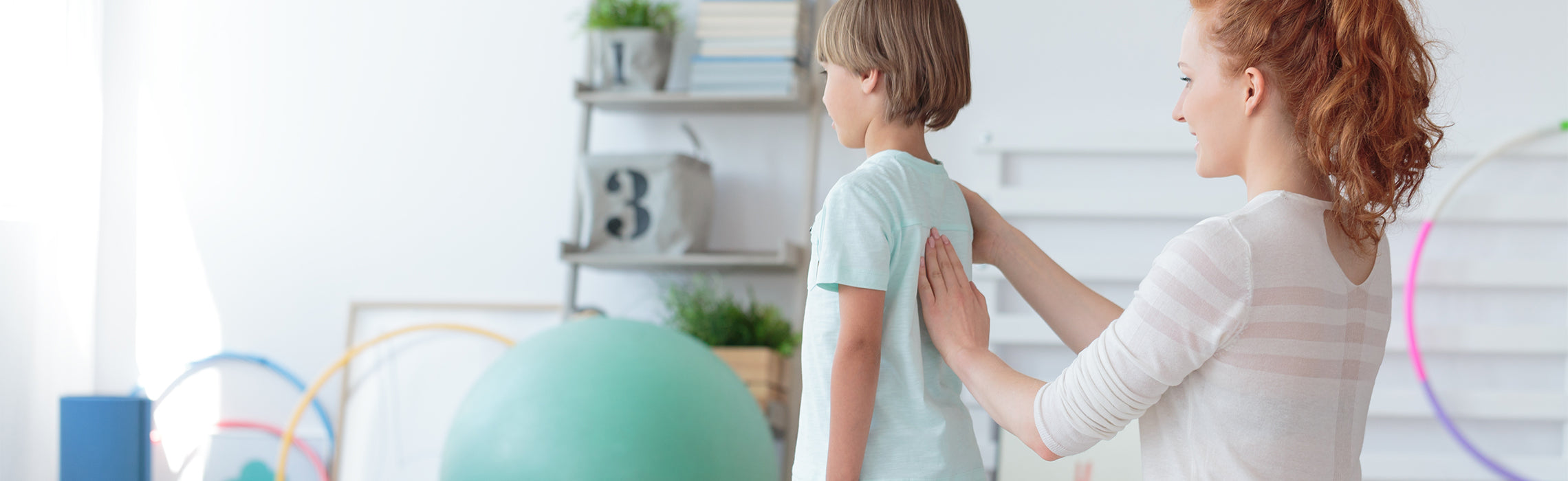 Healthcare provider placing a hand on a child’s upper back to assess posture in a bright therapy room with an exercise ball and hoops, representing pediatric posture evaluation, physical therapy, and child wellness.