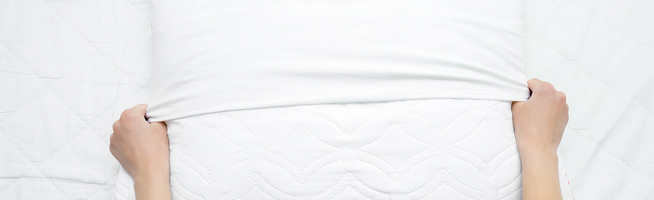 Hands pulling a crisp white fitted sheet over a mattress with a subtle quilted pattern, demonstrating bed-making or bedding care on a clean, minimal background. No visible text in the image.