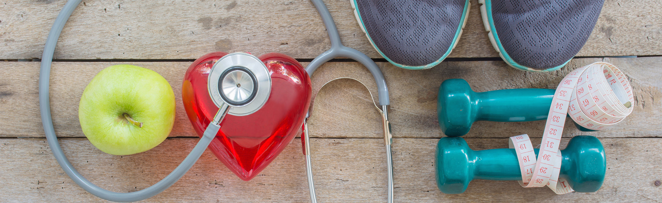 Flat lay of a green apple, red heart-shaped container with a stethoscope, teal dumbbells, measuring tape, and running shoes on a wooden surface, representing heart health, fitness, nutrition, and healthy lifestyle habits.