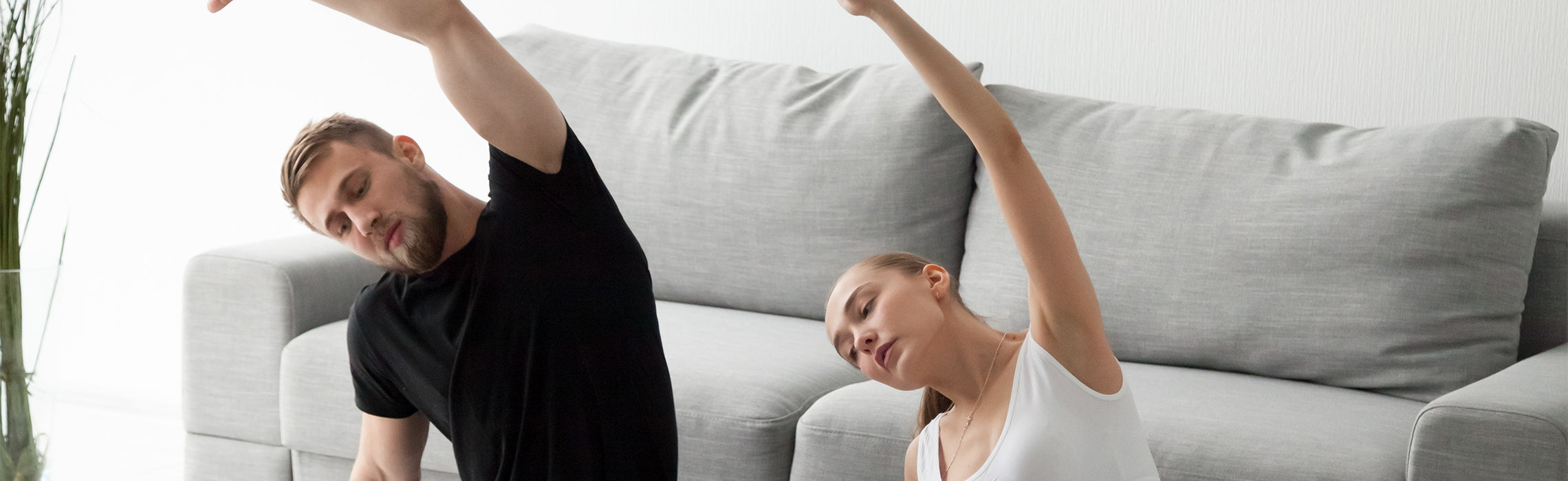 Two people performing side stretch exercises on a living room floor beside a gray sofa, raising one arm overhead in a home workout setting. No visible text or branding.