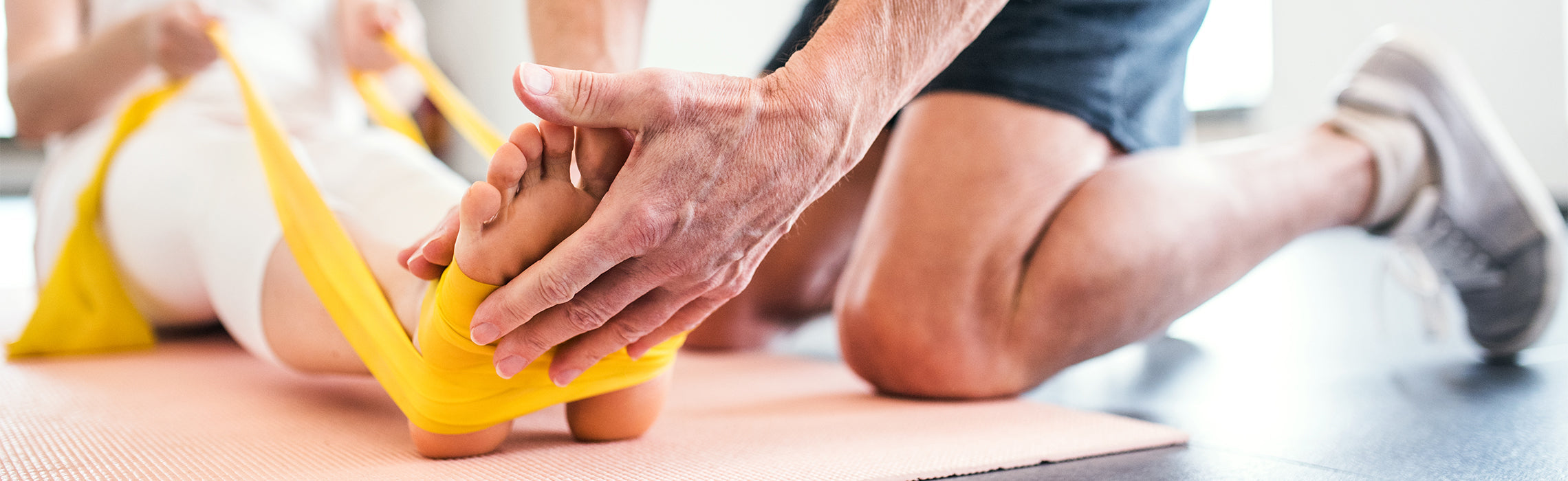 Close-up of a foot wrapped with a yellow resistance band while another person supports and stretches it on a yoga mat, demonstrating assisted foot rehabilitation, physical therapy, and flexibility training.