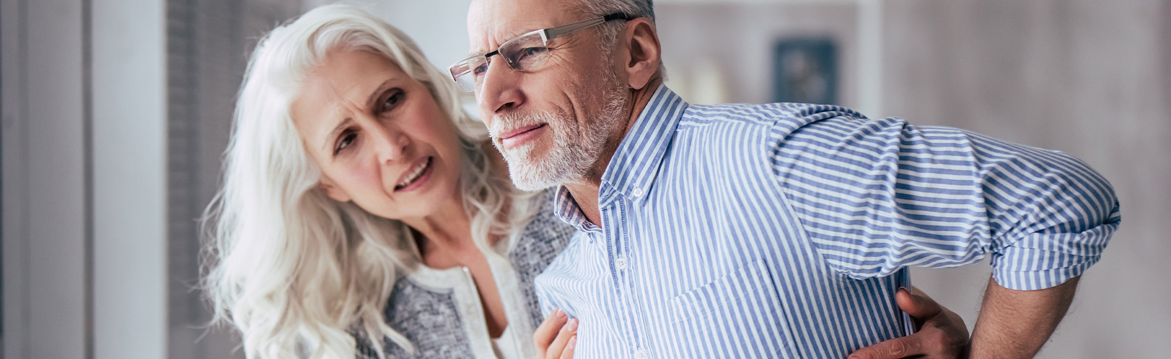 Senior man holding his lower back in discomfort while an older woman supports him beside a window indoors.