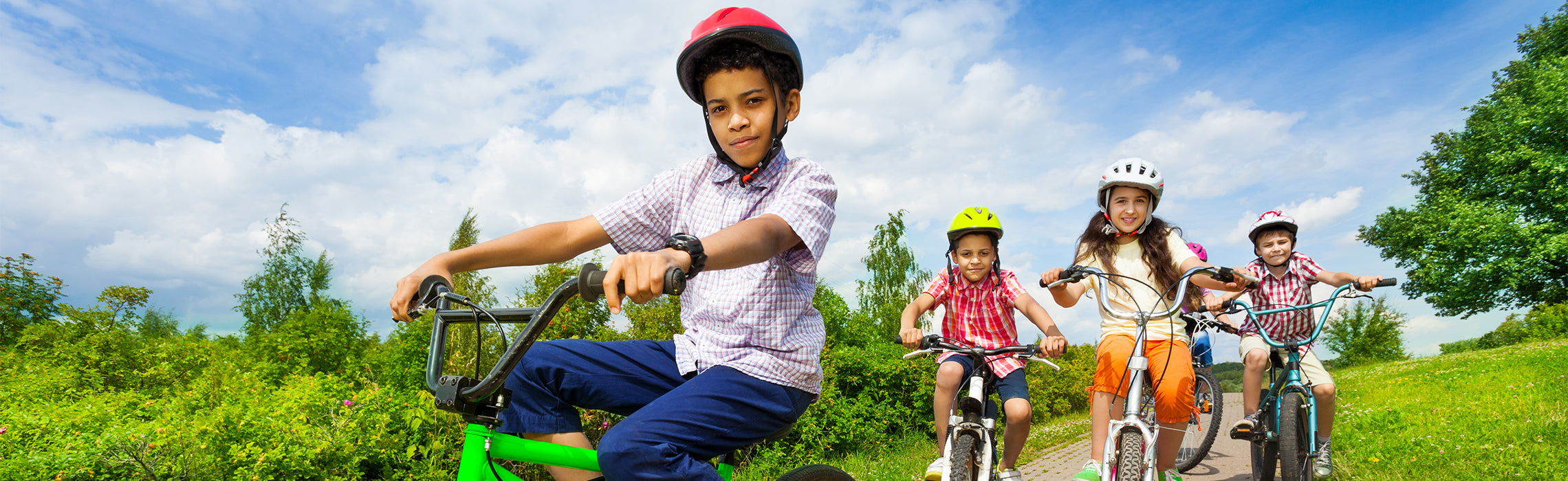 Four children riding bicycles on a paved path outdoors, all wearing safety helmets, with green grass, trees, and a blue sky in the background, representing kids’ outdoor activity, safety, and healthy lifestyle.
