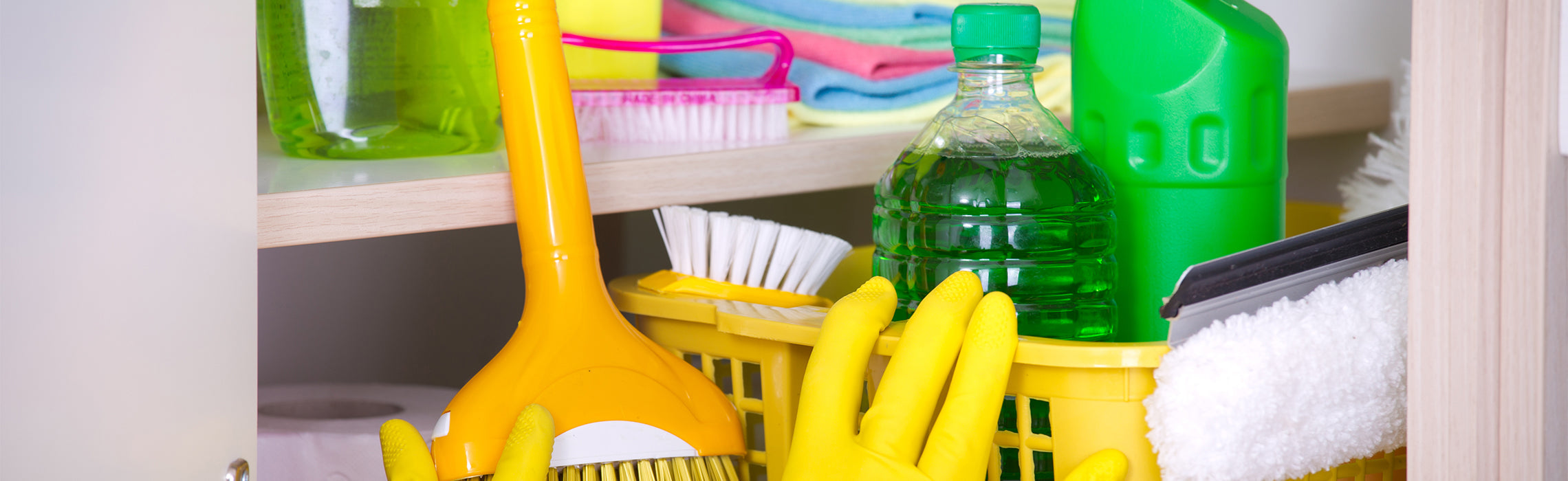 Cleaning supplies stored under a sink, including yellow rubber gloves, a brush, spray bottles, folded cloths, and green liquid cleaner in a plastic container. No visible text or branding.