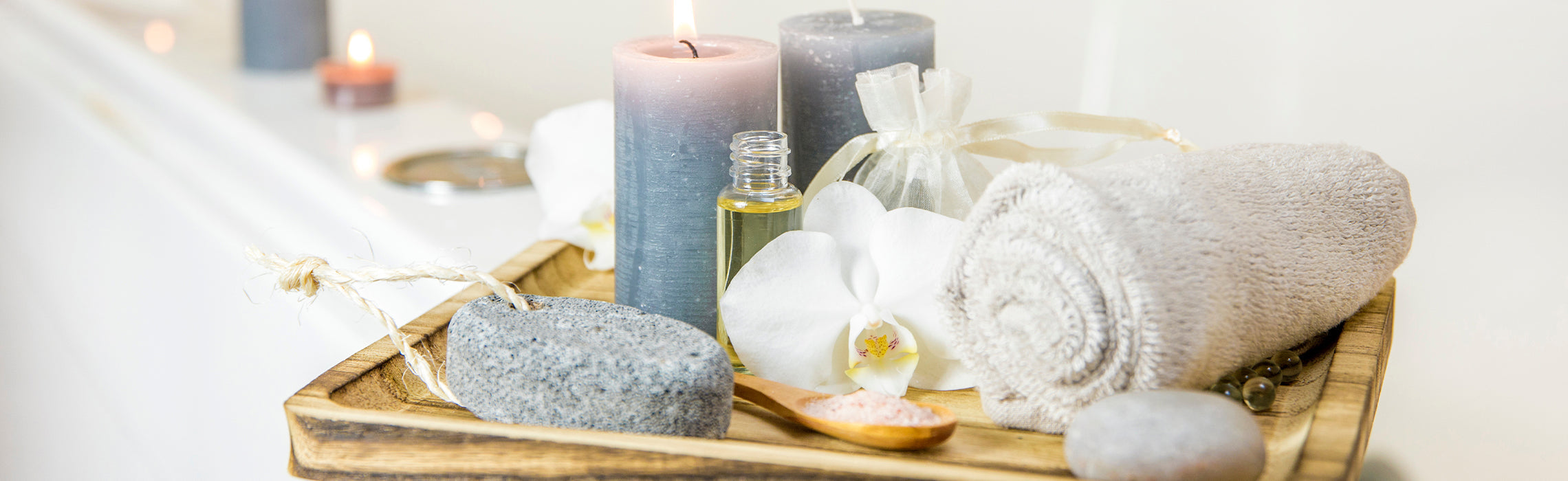 Spa setup with rolled towel, lit candles, essential oil bottle, bath salts, smooth stones, and a white orchid on a wooden tray, representing relaxation, wellness, and self-care.