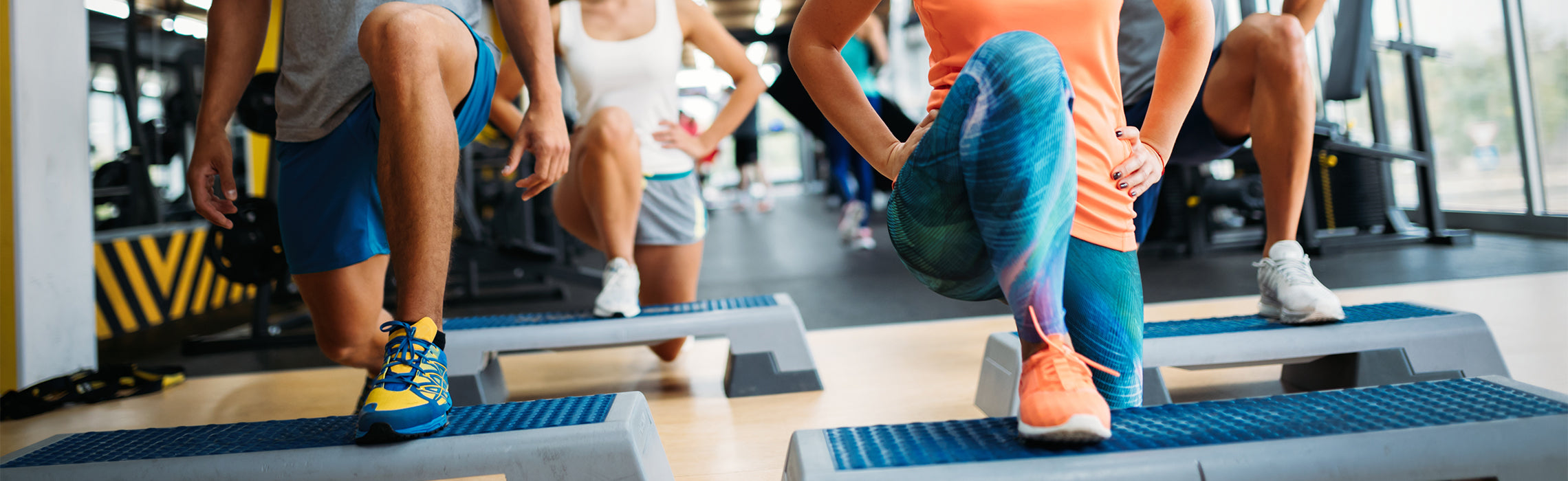 Group fitness class performing step-up lunges on aerobic platforms in a gym, with participants wearing athletic clothing and sneakers, representing cardio workout, strength training, and active lifestyle.