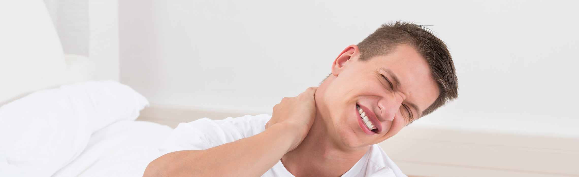 Close-up of a person sitting on a bed holding the back of the neck with one hand, with a pillow and bedding in the background, representing neck pain, stiffness, and sleep-related discomfort.