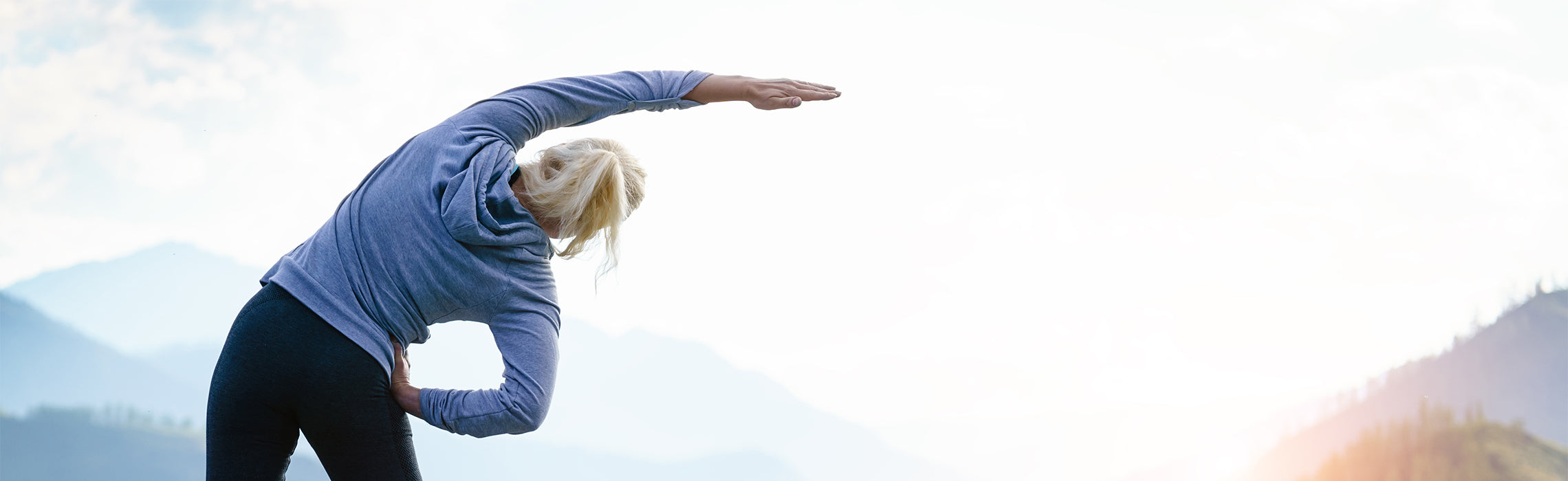 Person outdoors performing a side stretch with one arm overhead against a scenic mountain backdrop, representing flexibility exercise, outdoor fitness, and healthy active lifestyle.