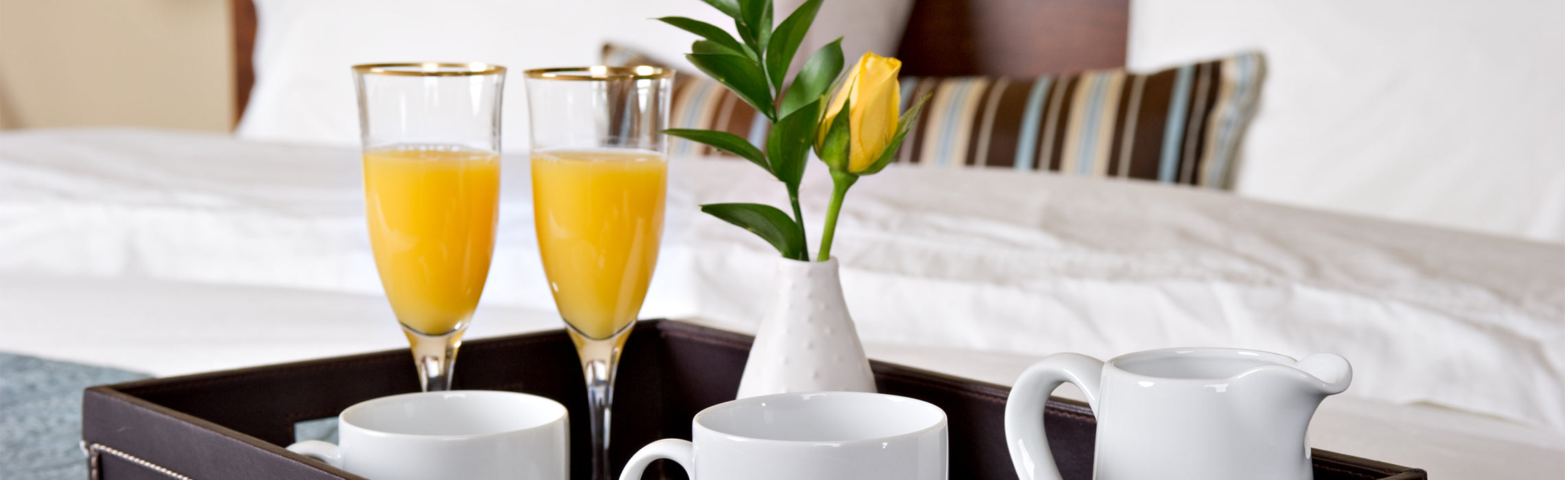 Breakfast in bed setup featuring orange juice, coffee cups, a teapot, and fresh flowers arranged on a serving tray atop white bedding.