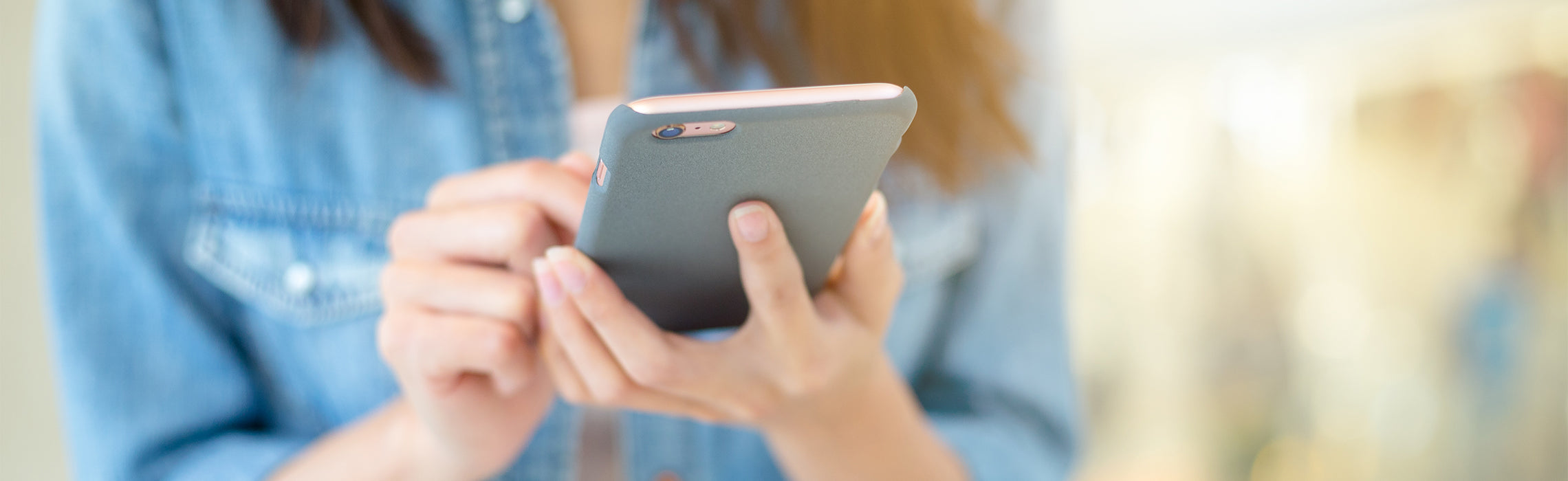 Close-up of a person using a smartphone with both hands, wearing a denim shirt in a softly lit indoor setting. No visible text or branding.