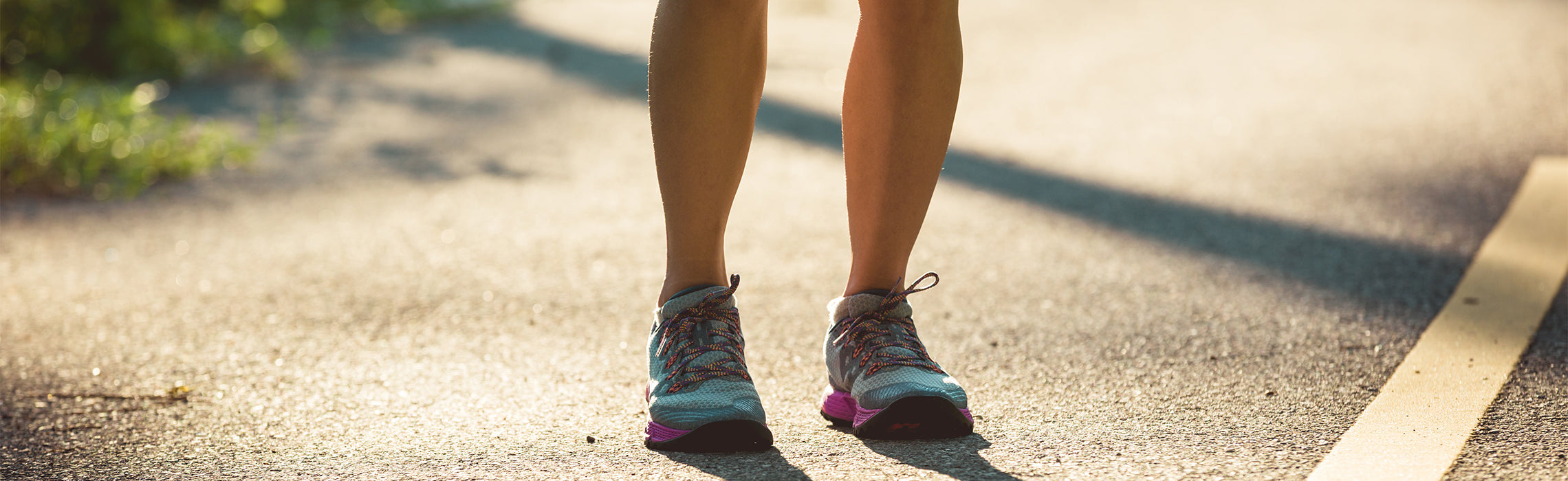 Close-up of a person’s legs and running shoes walking on a sunlit paved path outdoors.