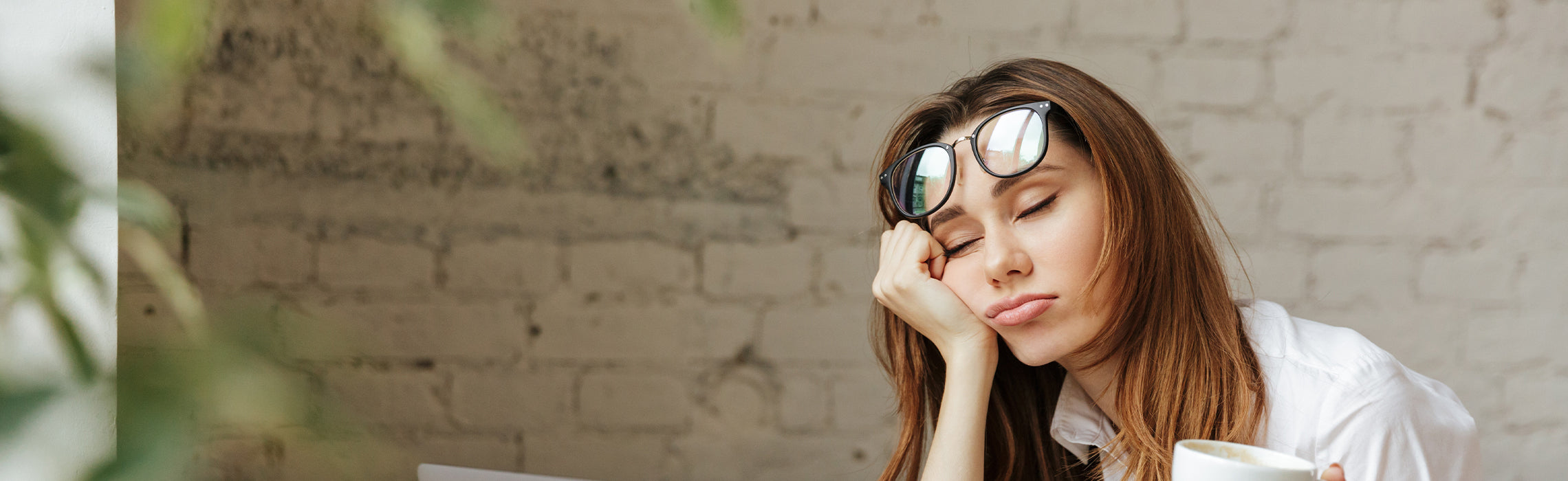 Close-up of a person with glasses resting on top of the head, leaning the face into one hand with eyes closed while seated indoors, holding a coffee cup, representing fatigue, eye strain, and workplace tiredness.