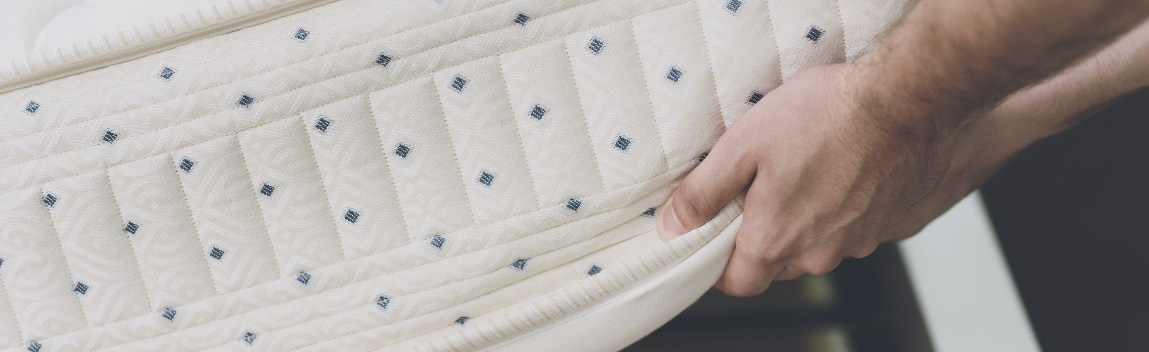 Close-up of hands lifting the edge of a quilted white mattress, showing layered construction and stitching detail, representing mattress durability, comfort, and sleep support.
