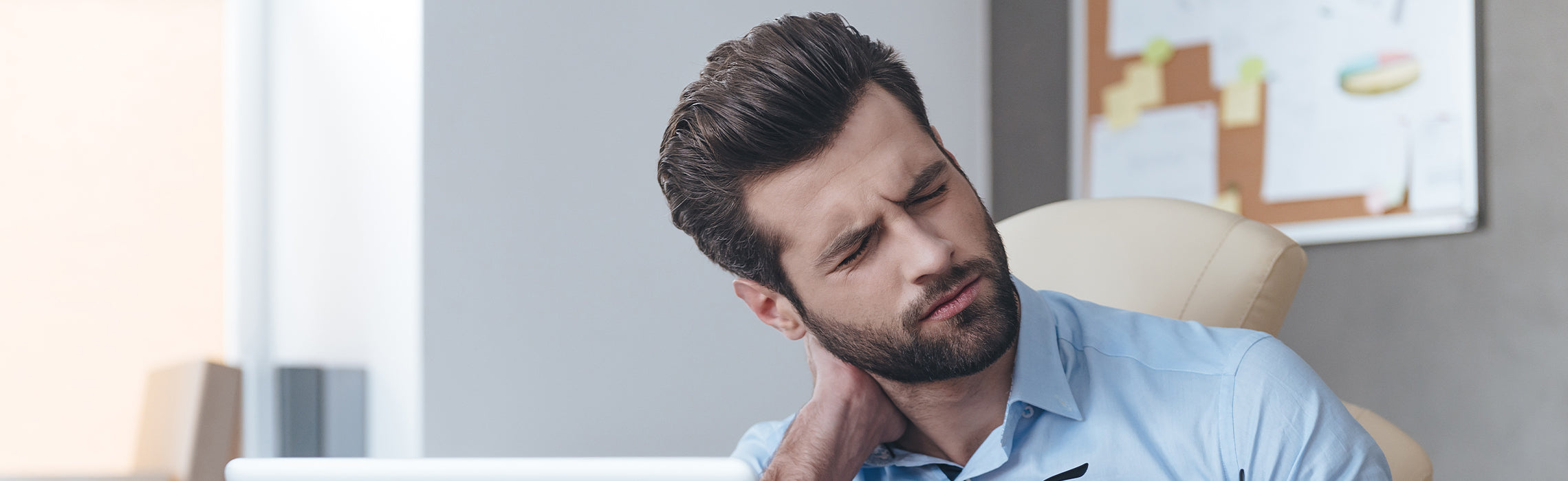 Close-up of a person seated at an office desk holding the back of the neck with one hand while tilting the head, with a laptop and office items nearby, representing neck pain, muscle strain, and work-related posture discomfort.