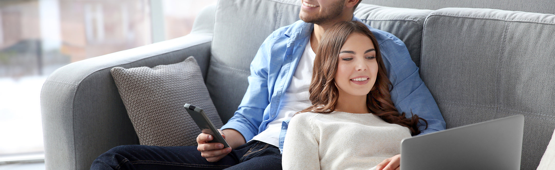Two people sitting on a couch using a laptop and smartphone in a bright living room, representing home technology use, online browsing, and a relaxed lifestyle.