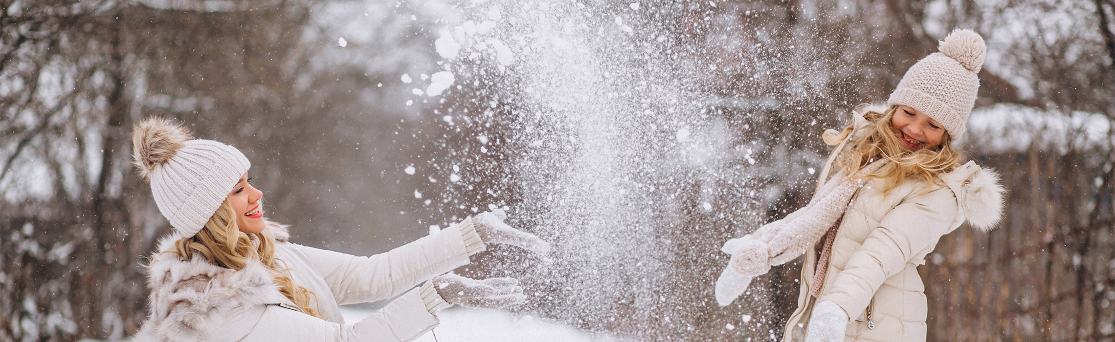 Two people outdoors in a snowy park tossing powdery snow into the air, wearing winter coats, knit hats, and gloves, with bare trees in the background, representing winter activities, seasonal fun, and outdoor recreation.