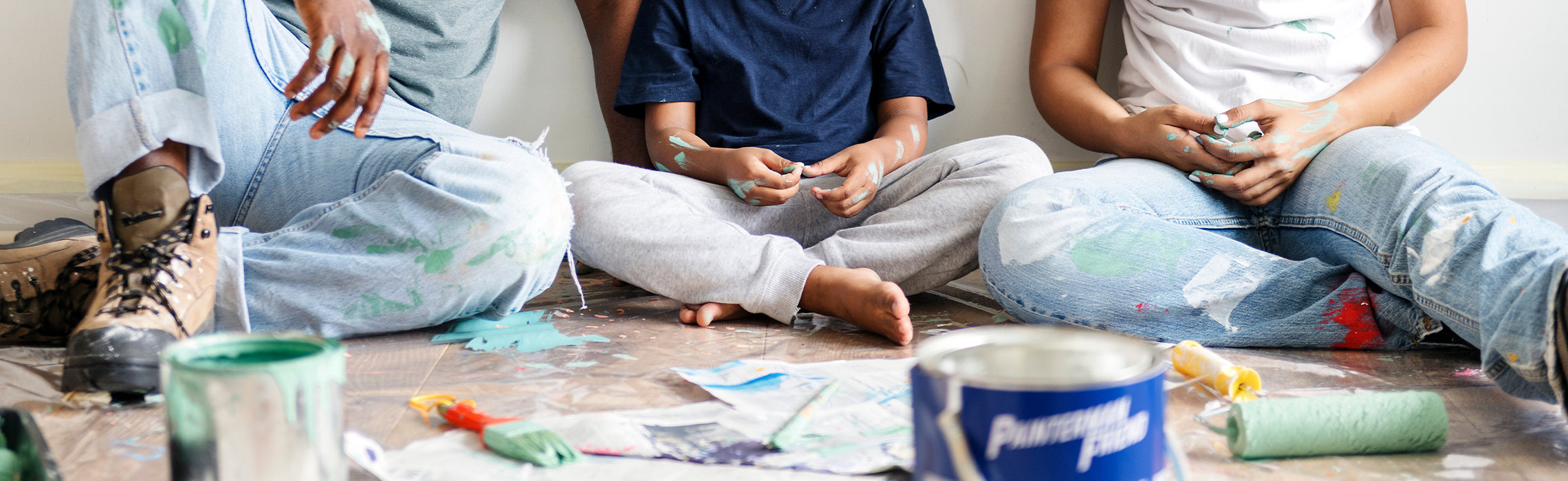 People sitting on a floor covered with a drop cloth, surrounded by paint cans, rollers, and brushes, with paint splatters on clothing and hands during a home painting project. No fully readable text or clear branding visible.
