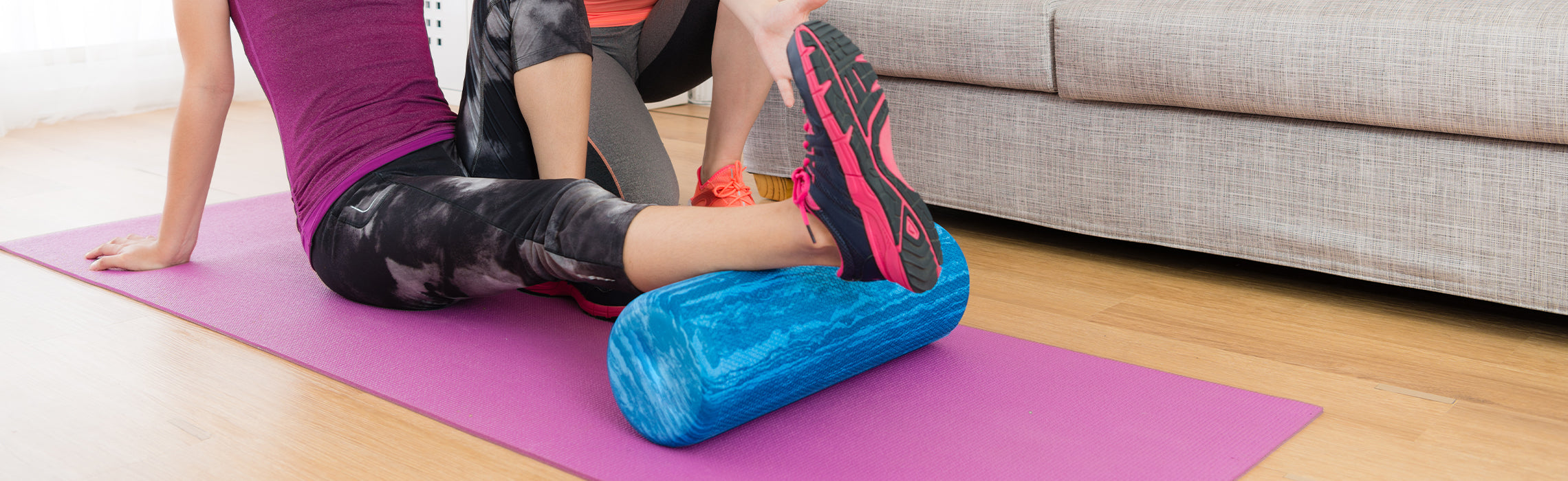 Person seated on a yoga mat using a blue foam roller under the leg while a trainer assists, demonstrating guided stretching, muscle recovery, and physical therapy exercises at home.