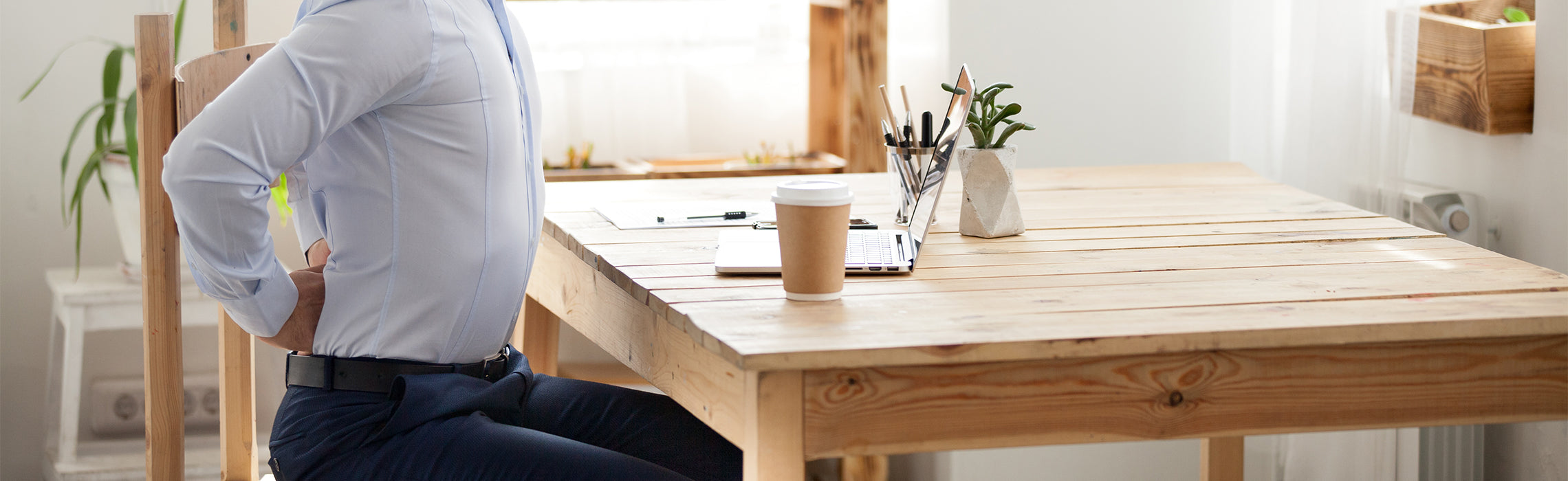 Person seated at a wooden desk holding the lower back with both hands near a laptop and coffee cup, representing back pain, poor posture, and workplace strain.