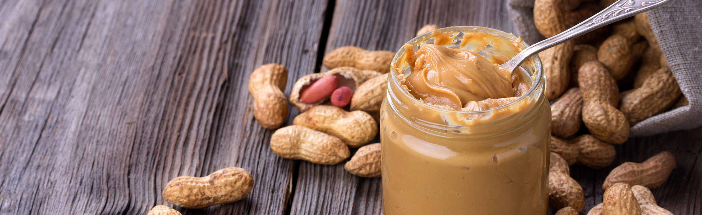 Open jar of creamy peanut butter with a spoon on a rustic wooden surface, surrounded by whole peanuts and a cloth bag, representing healthy snacks, protein-rich food, and natural nutrition.