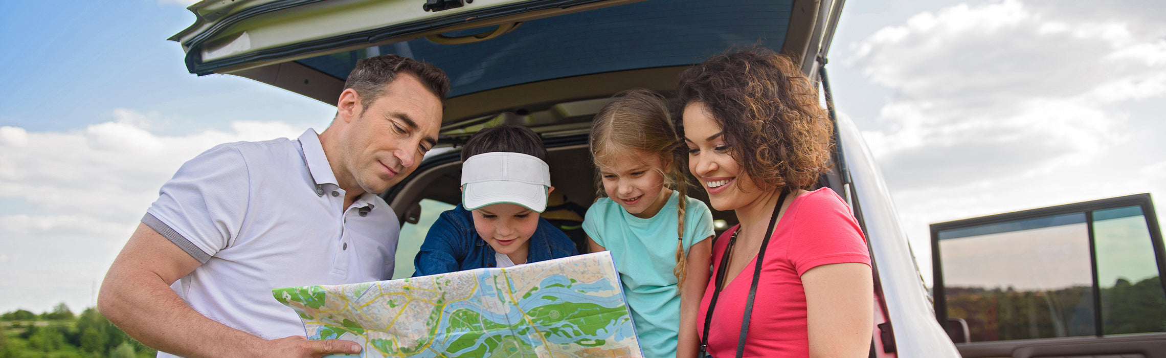 Family gathered at the open trunk of an SUV reviewing a map during a road trip, with adults and children looking at directions outdoors. No visible text or branding.